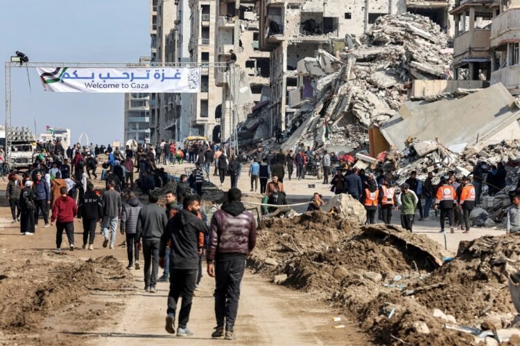 People gather by a banner welcoming people near the rubble of a collapsed building along Gaza's coastal al-Rashid Street for people to cross from the Israeli-blocked Netzarim corridor from the southern Gaza Strip into Gaza City on January 26, 2025. Israel said on January 25 it would block the return of displaced Palestinians to their homes in northern Gaza until Arbel Yehud, one of the hostages taken captive during the October 7, 2023 attacks, is released. Hamas sources said that Yehud was "alive and in good health", and would be "released as part of the third swap set for next Saturday", on February 1. (Photo by Omar AL-QATTAA / AFP)